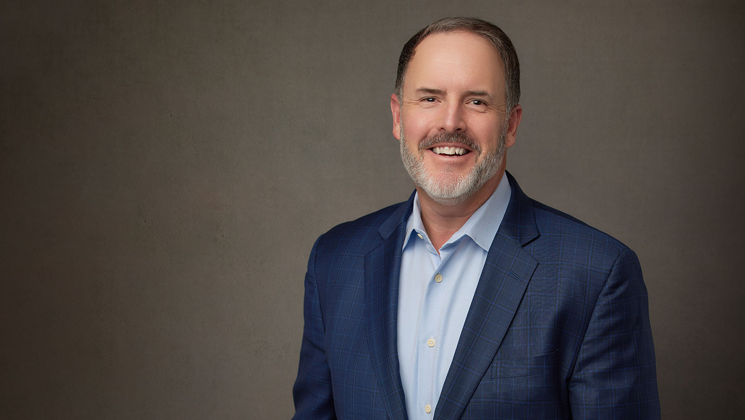 A white man with a beard in a blue shirt and sportcoat smiles against a textured brown background